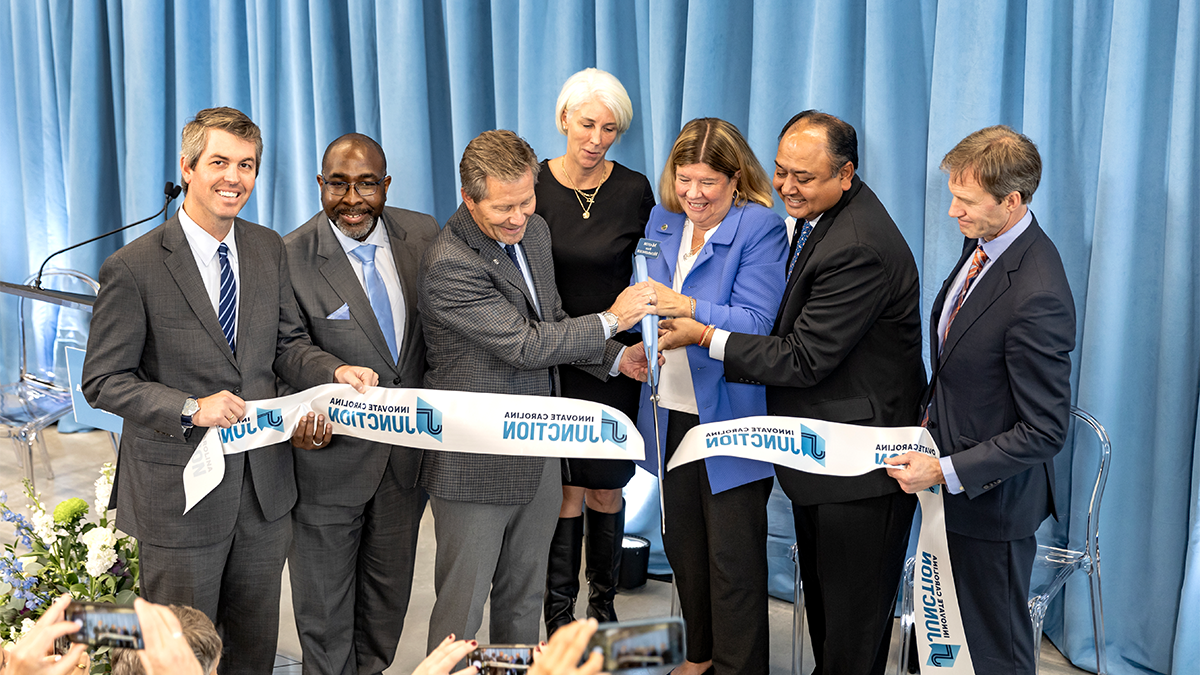 A group of six people standing in front of an audience and cutting a ribbon with a large pair of scissors at a ribbon-cutting event for the Innovate Carolina Junction.